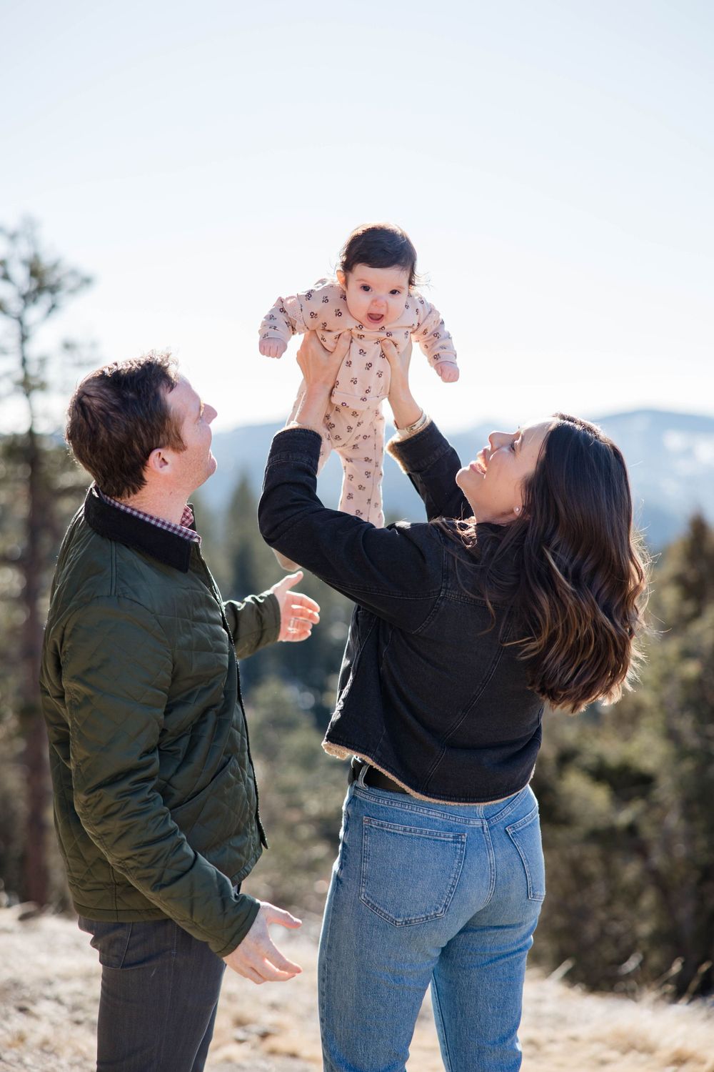 A joyful outdoor family photoshoot in nature with a small baby in a floral onesie being lifted in the air.
