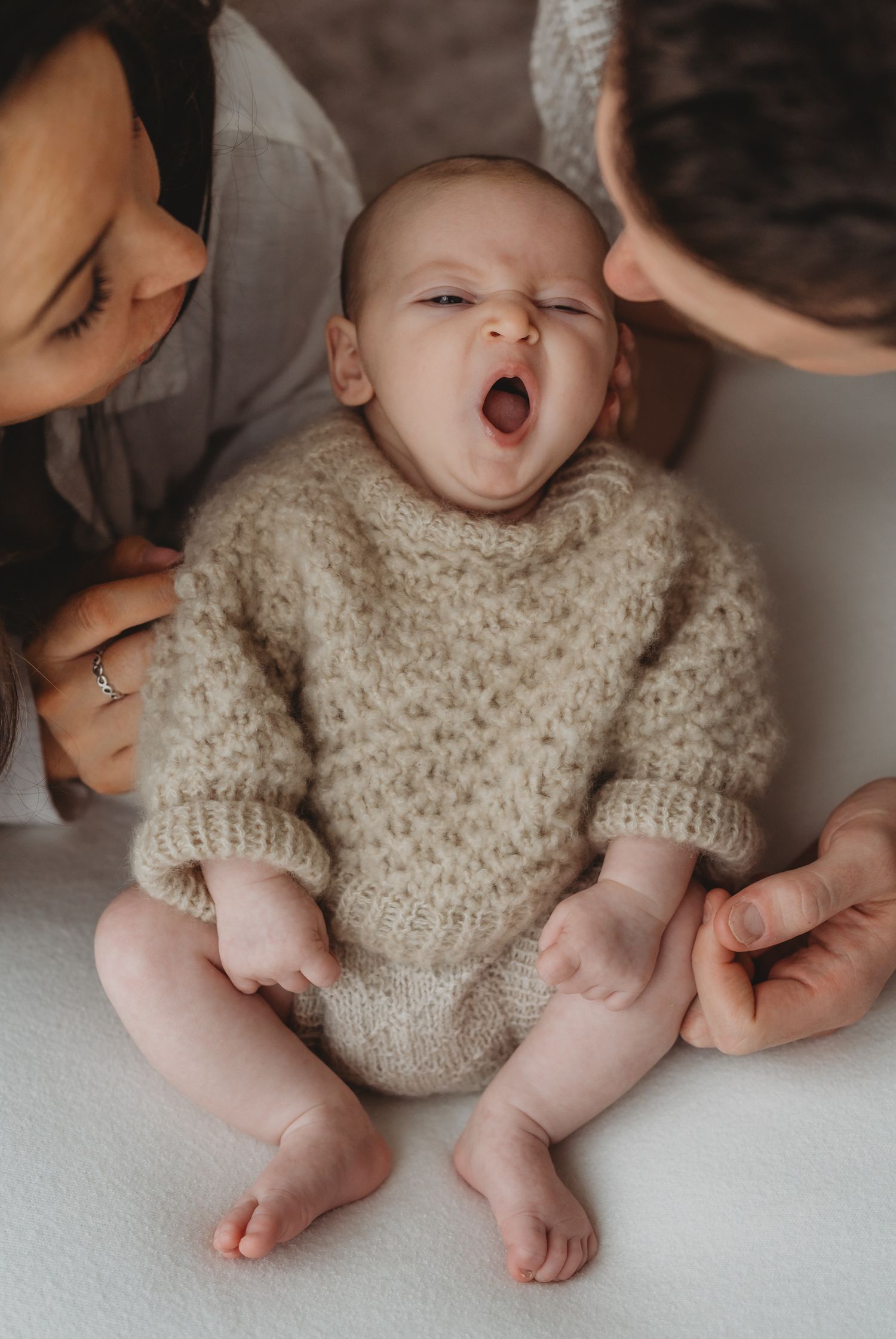 Leicestershire newborn photographer. Yawning baby wearing cozy knit sweater sits between parents in intimate lifestyle portrait.
