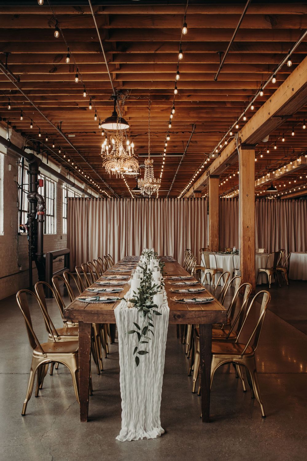 Rustic industrial wedding reception venue with wooden tables, gold chairs, macrame runner and crystal chandeliers.