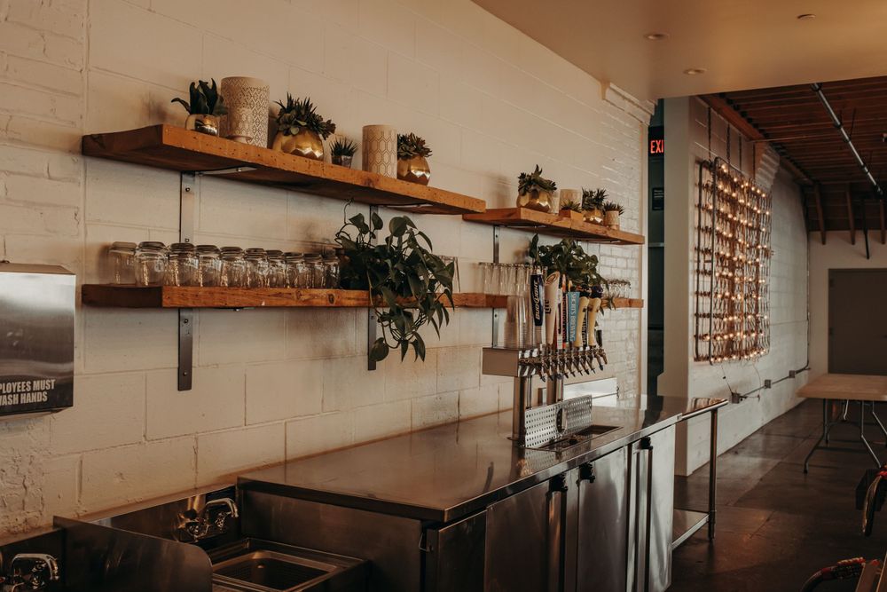 Rustic cafe interior with wooden shelves displaying potted plants and glassware against a white brick wall.
