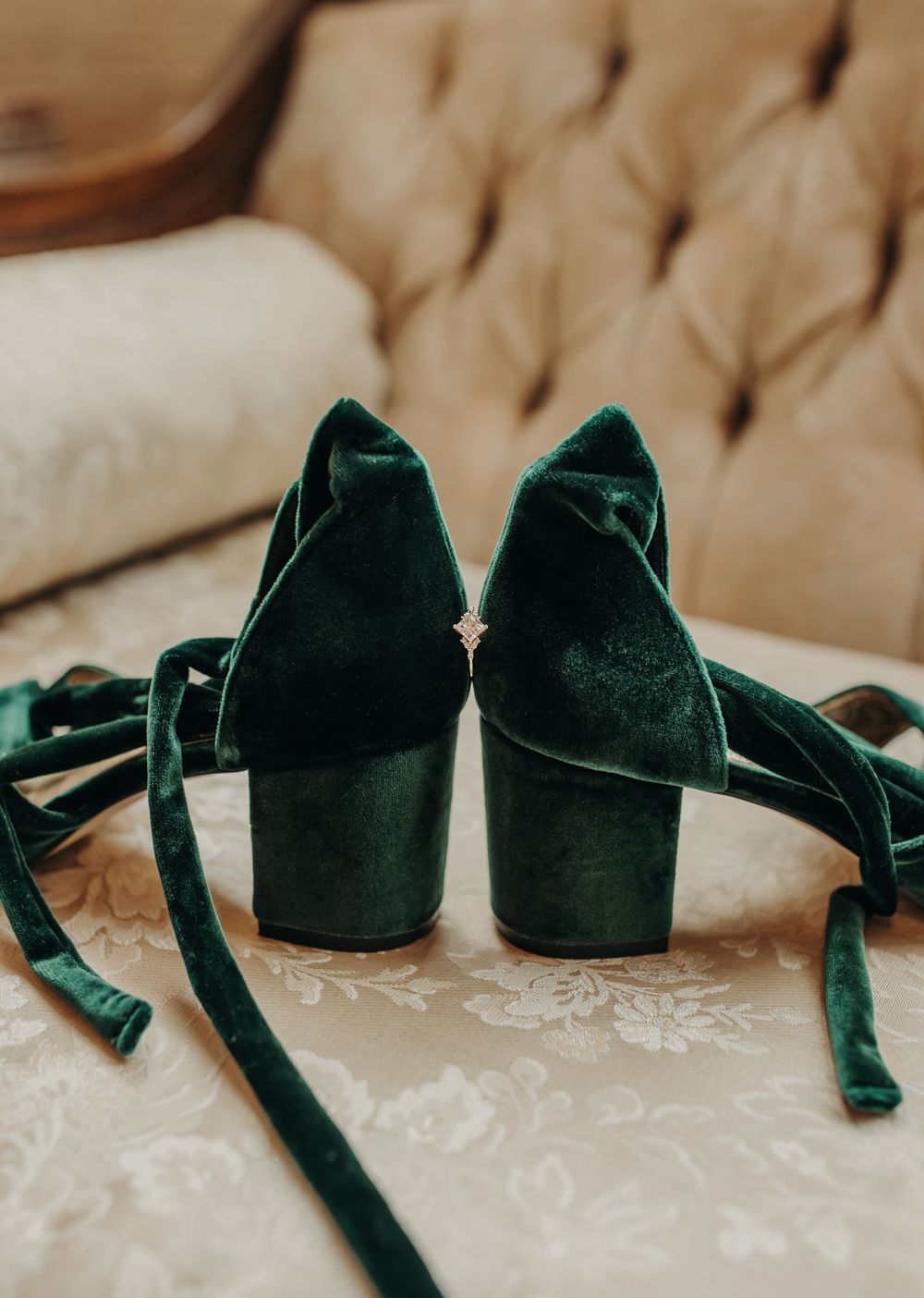 Emerald green velvet shoes with block heels and ankle straps rest on a cream-colored cushioned surface.