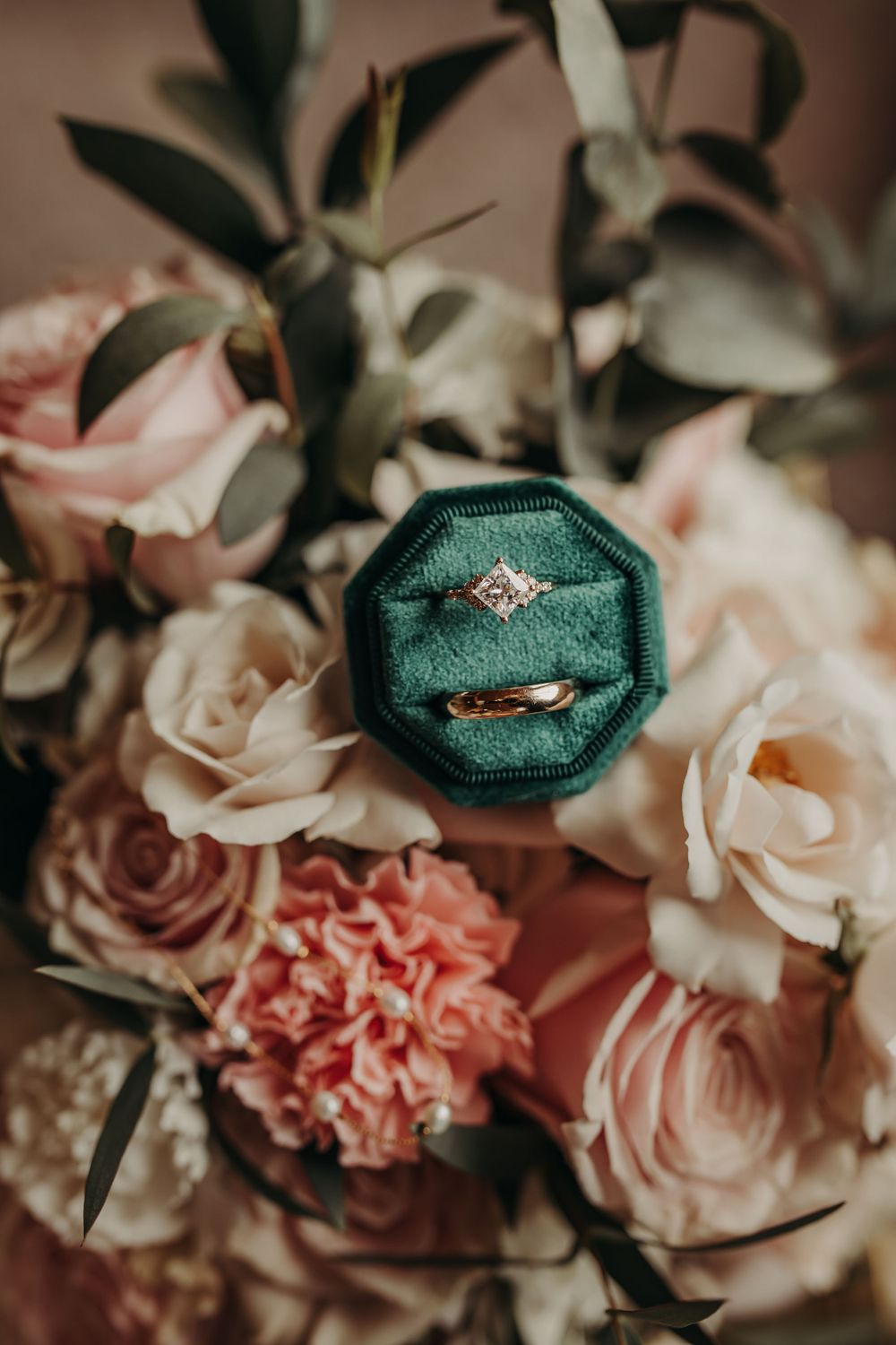 An engagement ring in emerald green velvet box sits atop blush pink and peach roses and carnations with greenery.