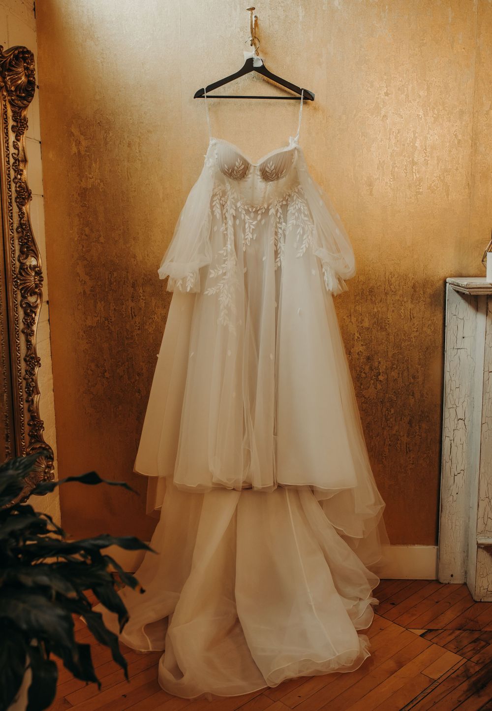 Elegant white wedding dress with sweetheart neckline and flowing tulle skirt hanging on wooden hanger against golden wall.