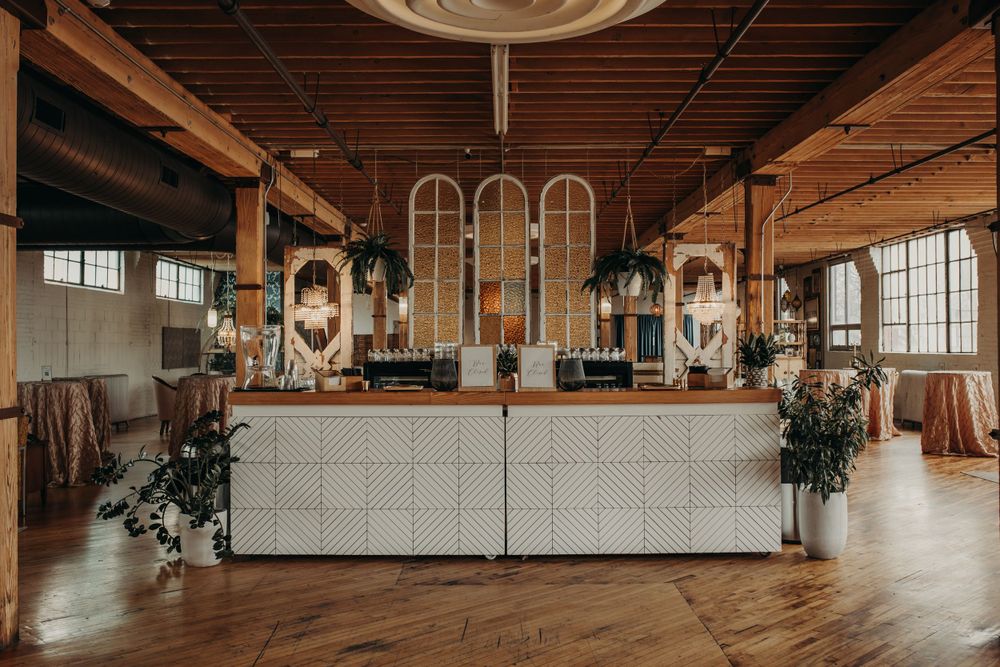 Rustic industrial wedding venue with white bar, exposed beams, arched windows, and floral arrangements.