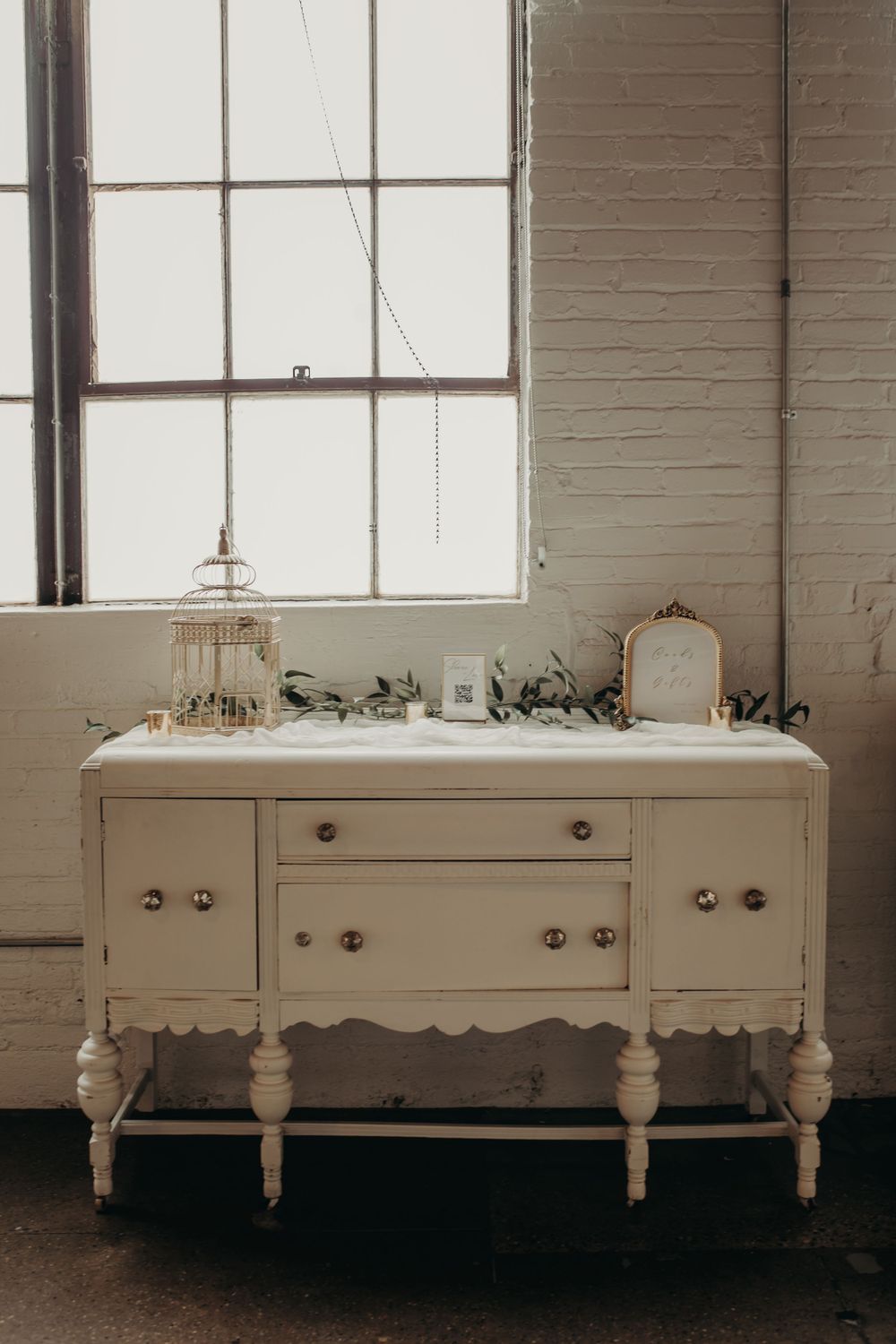 A vintage white painted sideboard with turned legs and brass hardware sits beneath a bright window in a rustic room.