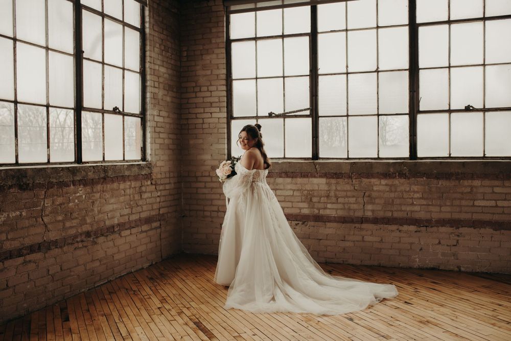 A bride in a flowing white gown stands near large industrial windows in a rustic brick-walled loft space with wooden floors.