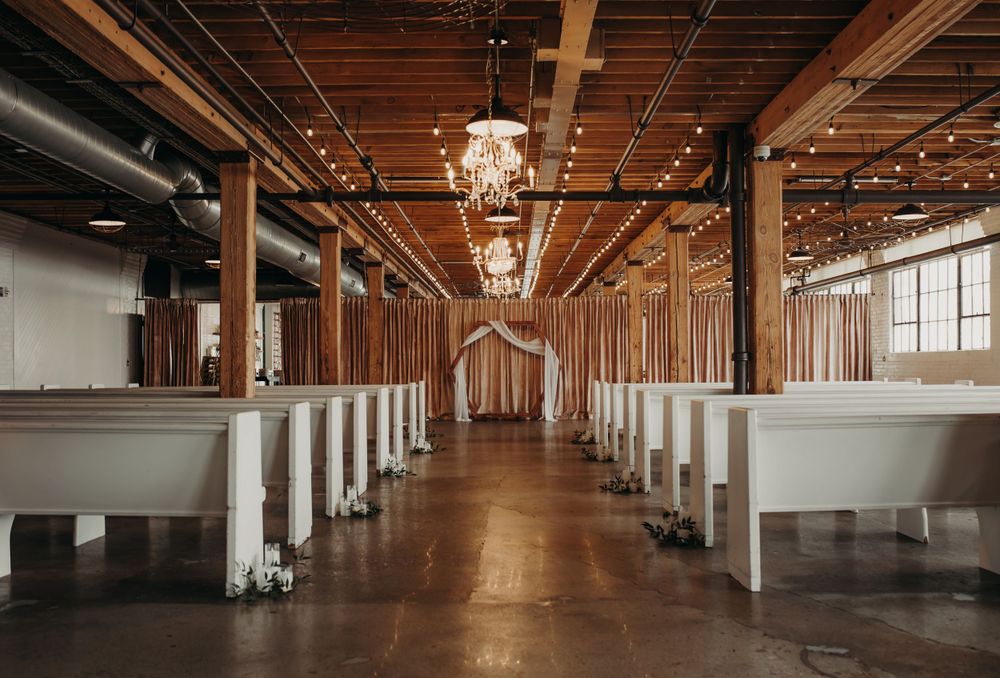 Elegant wedding venue interior with white benches, exposed wooden beams, and crystal chandeliers in a rustic-modern space.