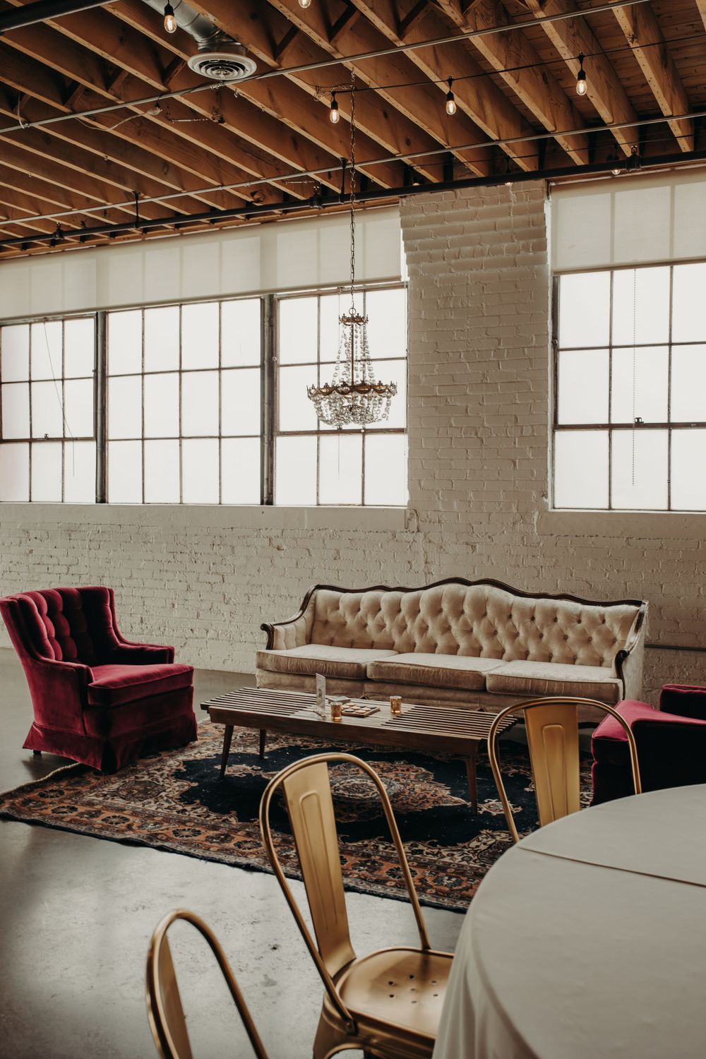 Industrial loft space with vintage furniture including tufted beige sofa, red velvet chairs, crystal chandelier and exposed beams.