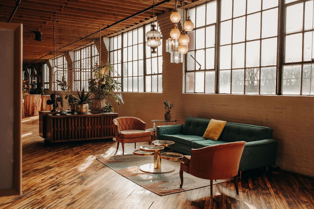 Warm industrial loft interior with emerald green sofa, vintage accent chairs and large factory windows with natural light.