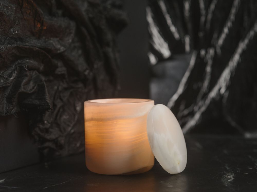 A glowing orange candle in a frosted glass jar with an open lid against a dark moody background.