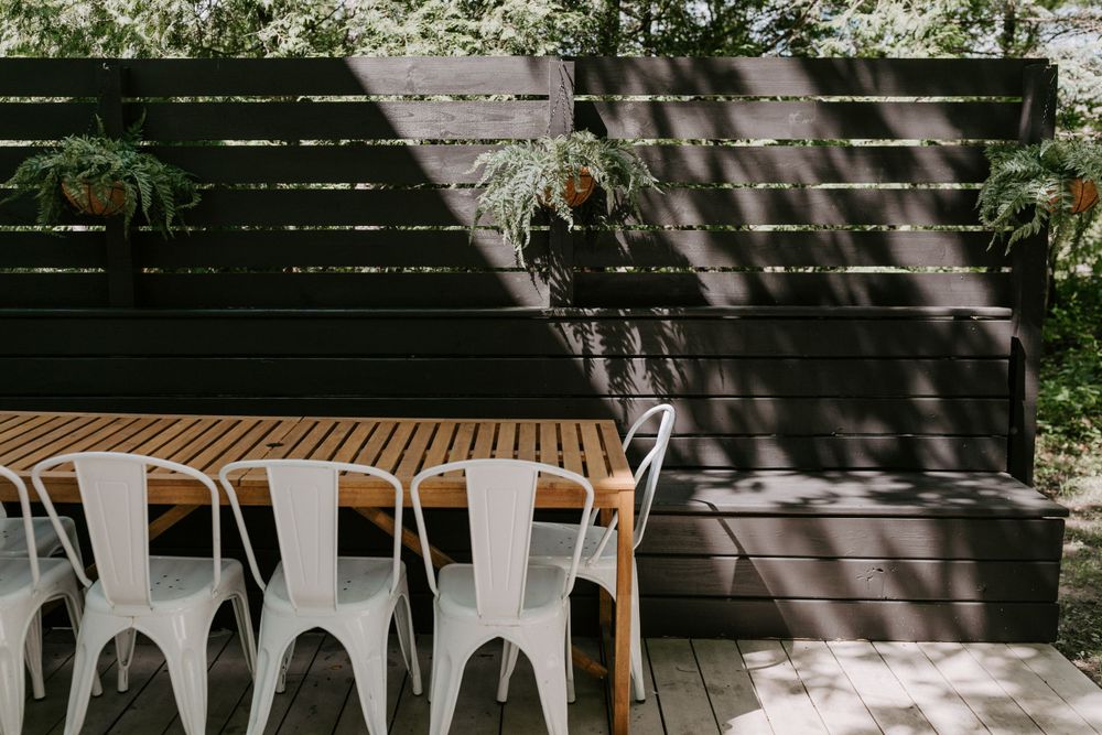 Modern outdoor dining area with white metal chairs and wooden table against a dark fence.