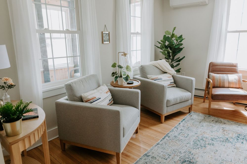 Modern living room with gray armchairs, leather accent chair, and houseplants near large windows.