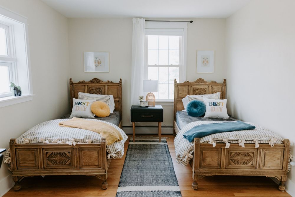 Two vintage wooden twin beds with decorative pillows flank a small nightstand in a bright white bedroom.