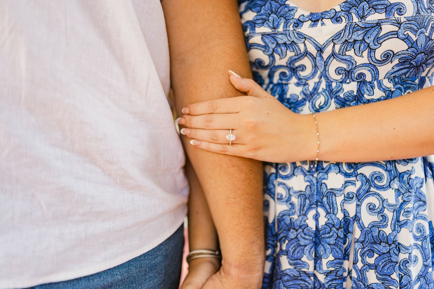 Couple embracing, woman wearing engagement ring and bracelet in blue floral dress beside man in white shirt.
