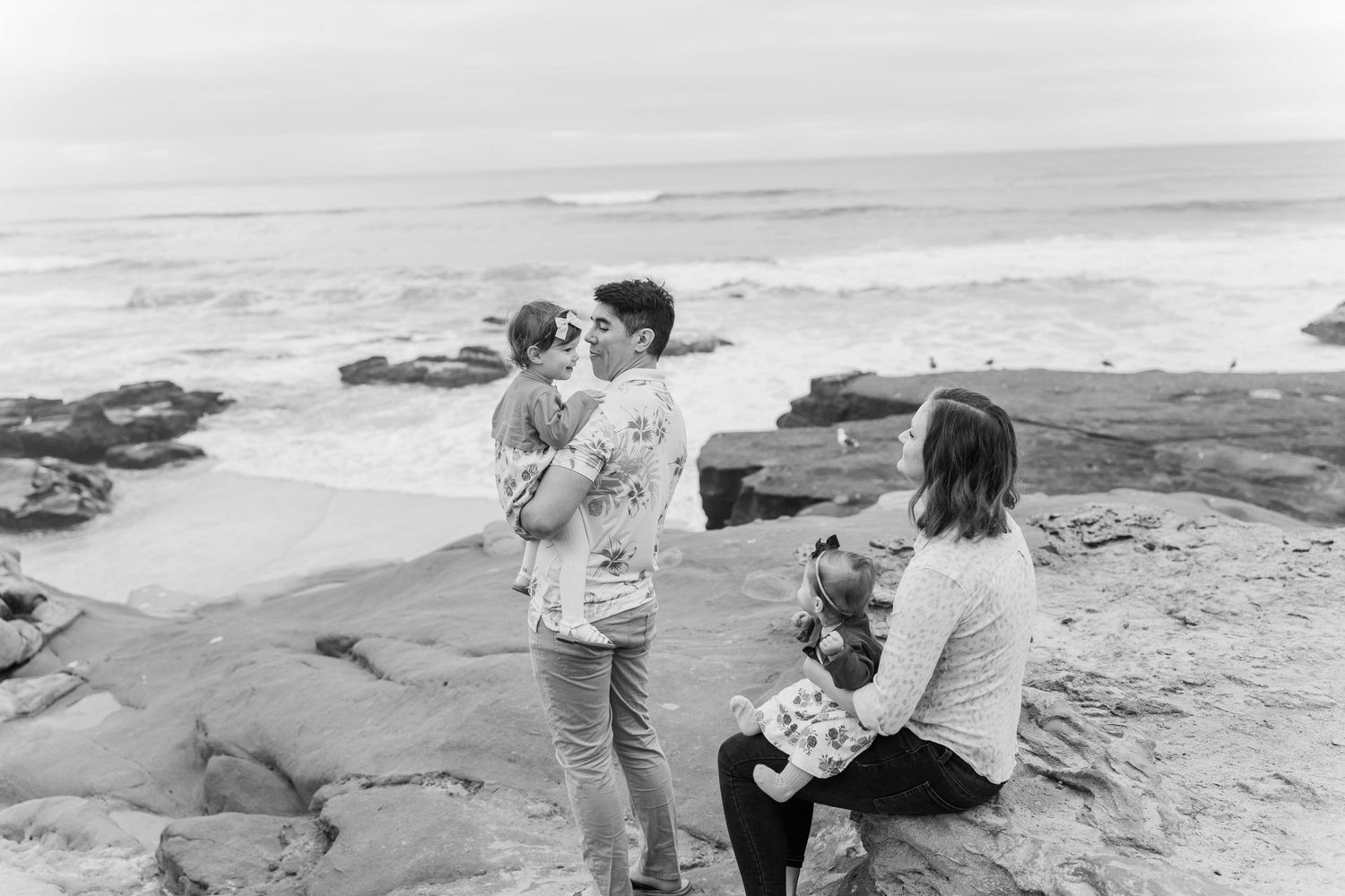 Family bonding moment captured on rocky beach with crashing waves in background.