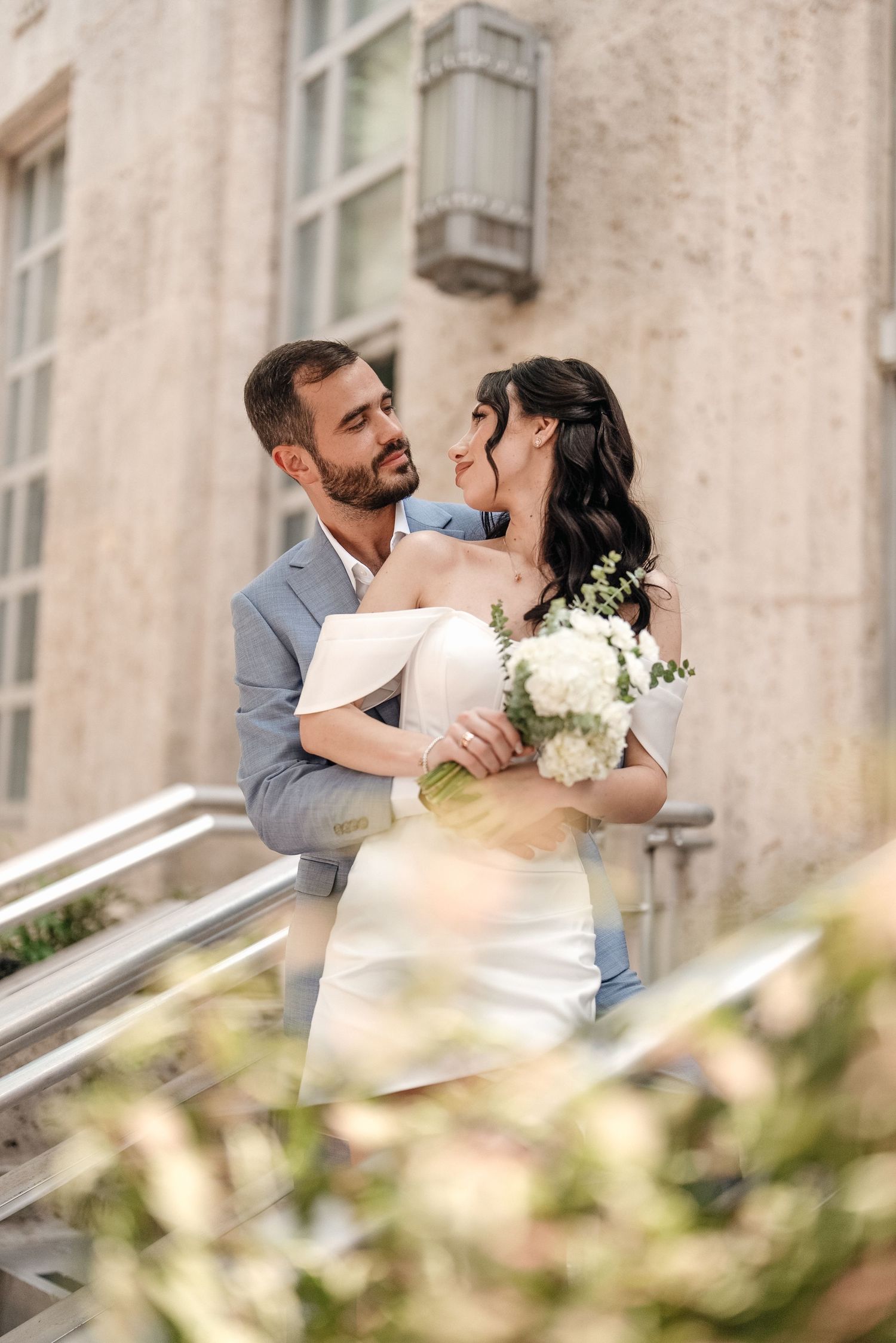 A romantic wedding moment against a light stone wall showing the couple embracing with white flowers.