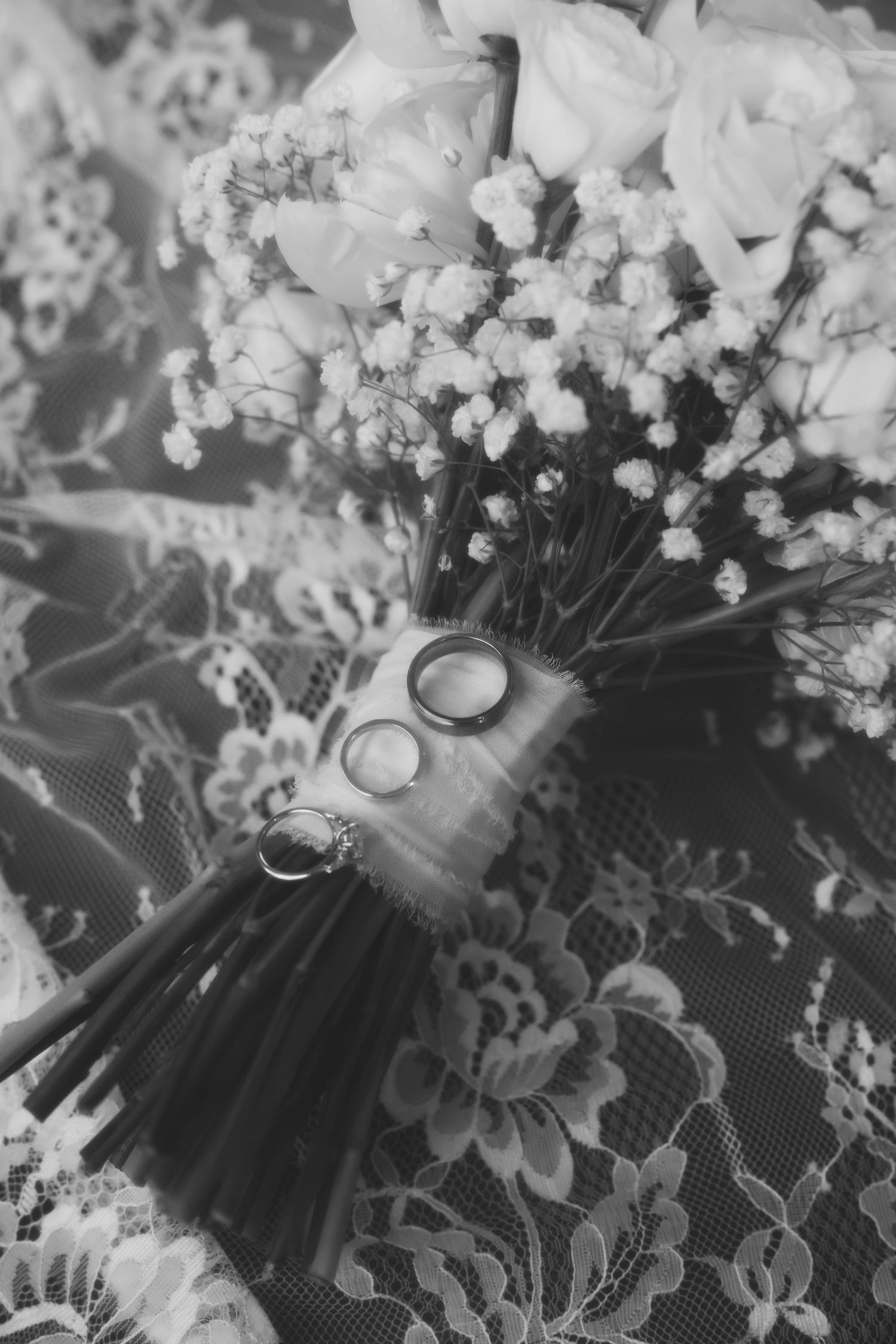 Black and white photo of wedding rings resting on a bridal bouquet with roses and baby's breath on lace fabric.