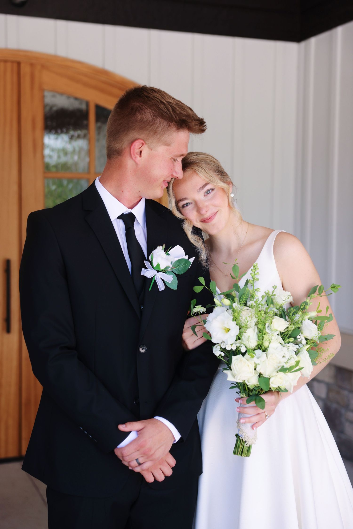 A bride and groom pose together, she holds white florals, he wears a black suit with boutonniere, outdoors near wooden doors.