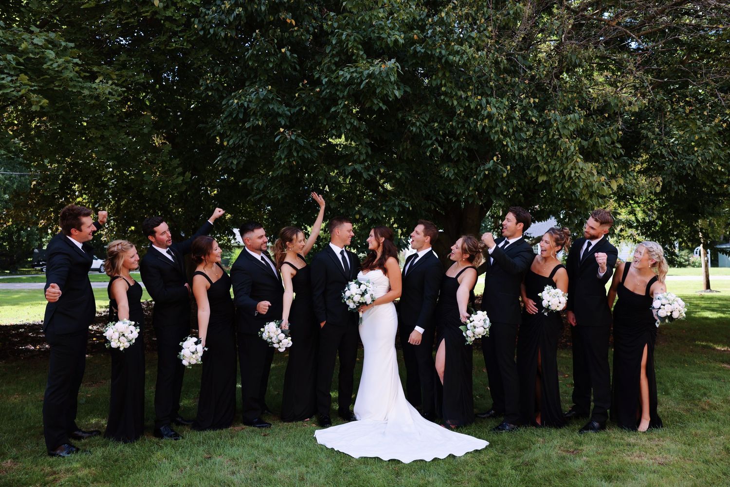 Wedding party in black attire gathers around bride in white dress for formal outdoor photo on green lawn.