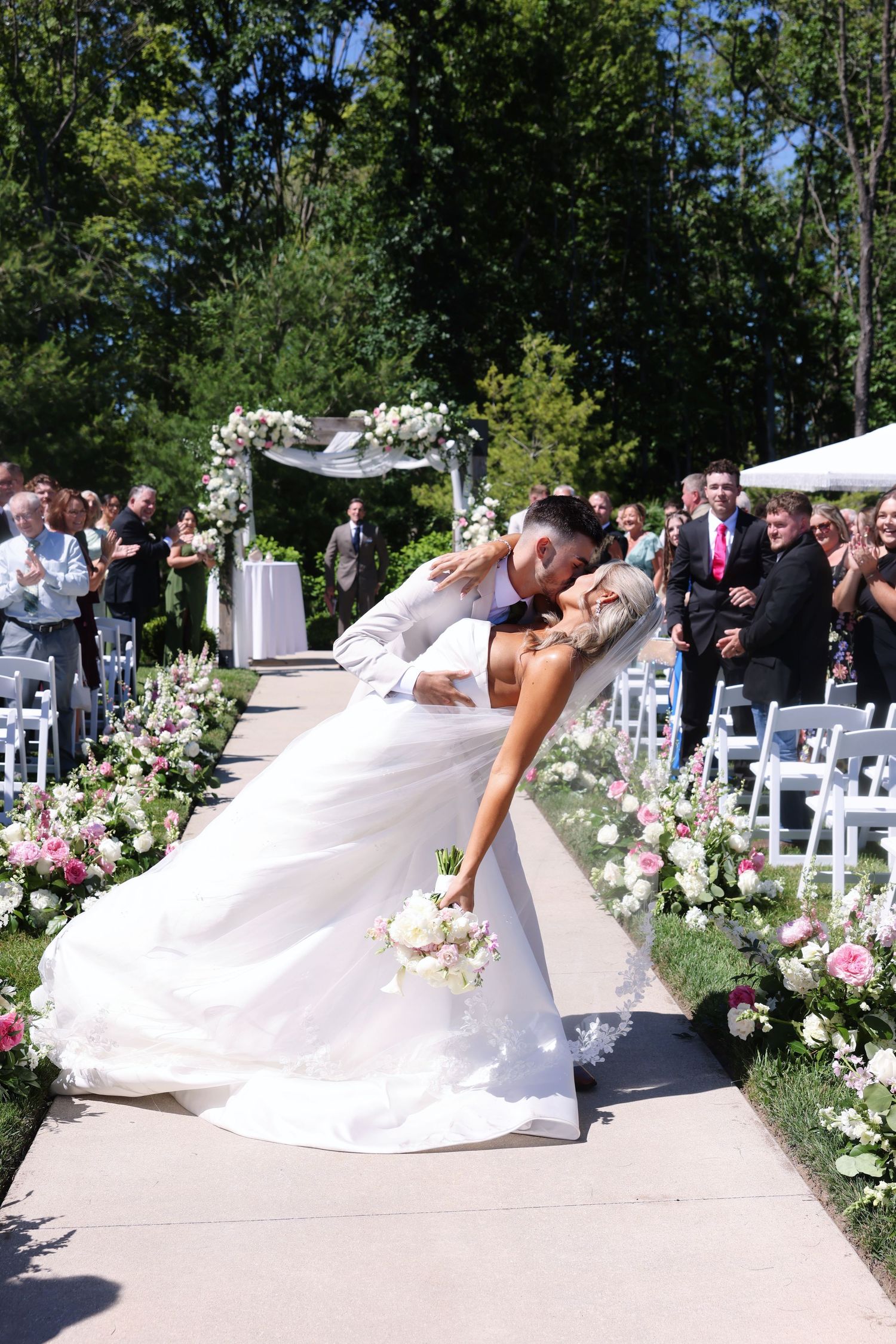 Bride and groom share a romantic dip kiss at outdoor wedding ceremony aisle lined with pink flowers.