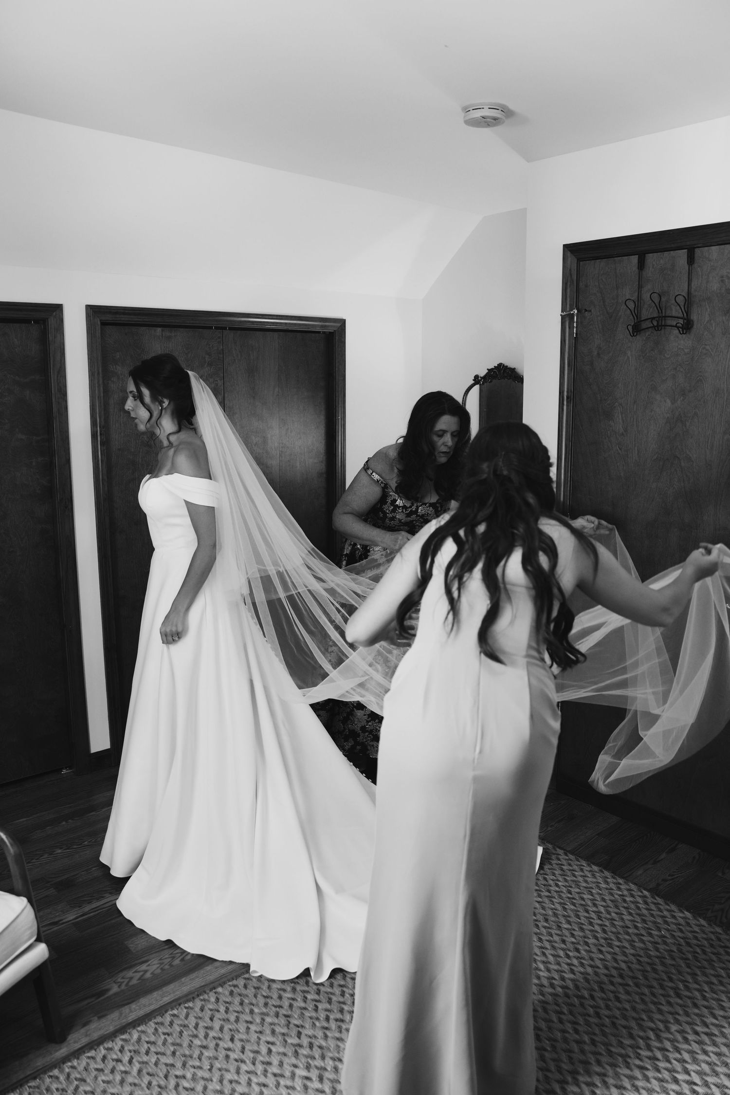 Behind-the-scenes wedding preparation moment showing bridal party members helping with dress and veil in black and white.