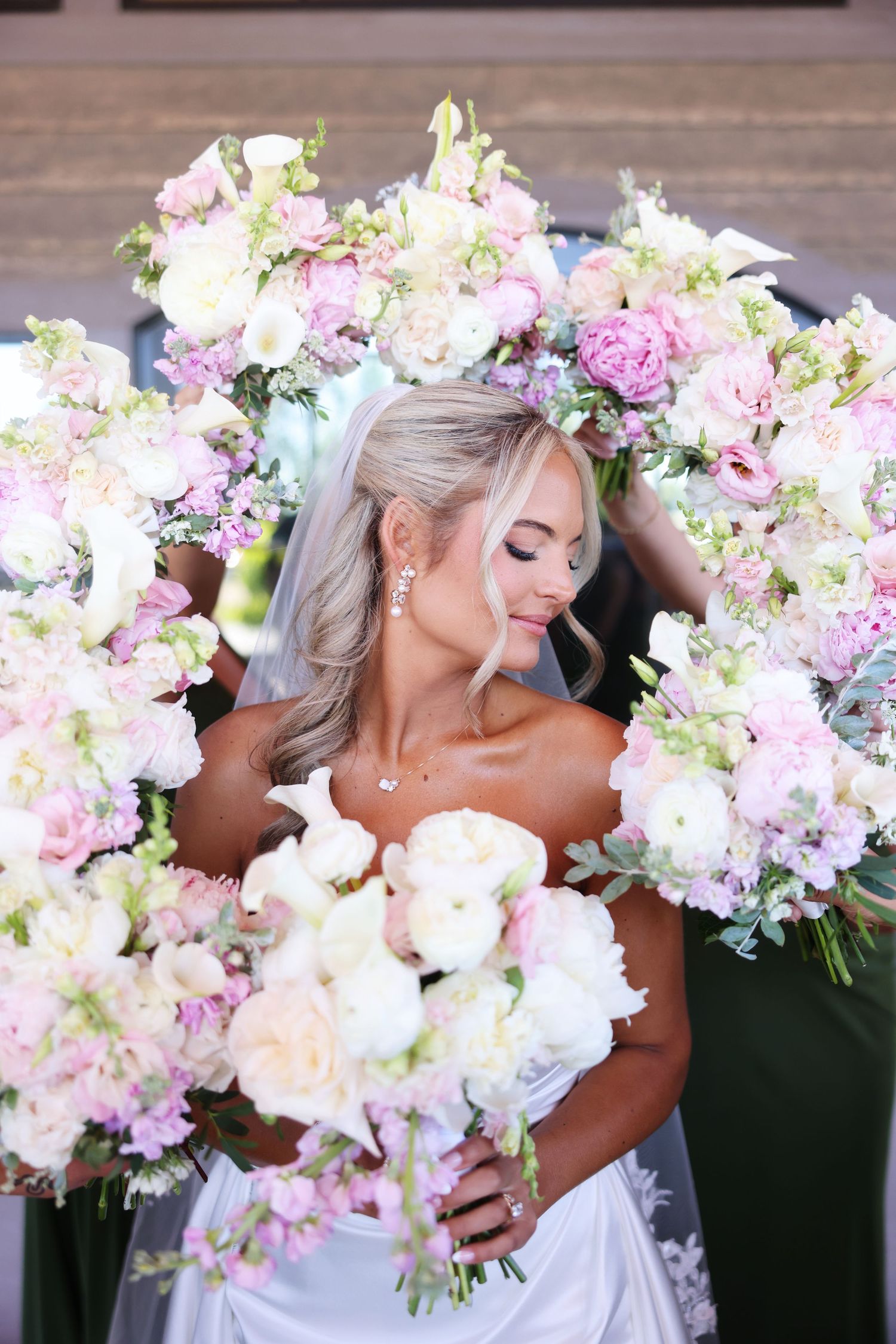 Bride in white gown surrounded by lush pink and white floral bouquets, smiling softly on her wedding day.