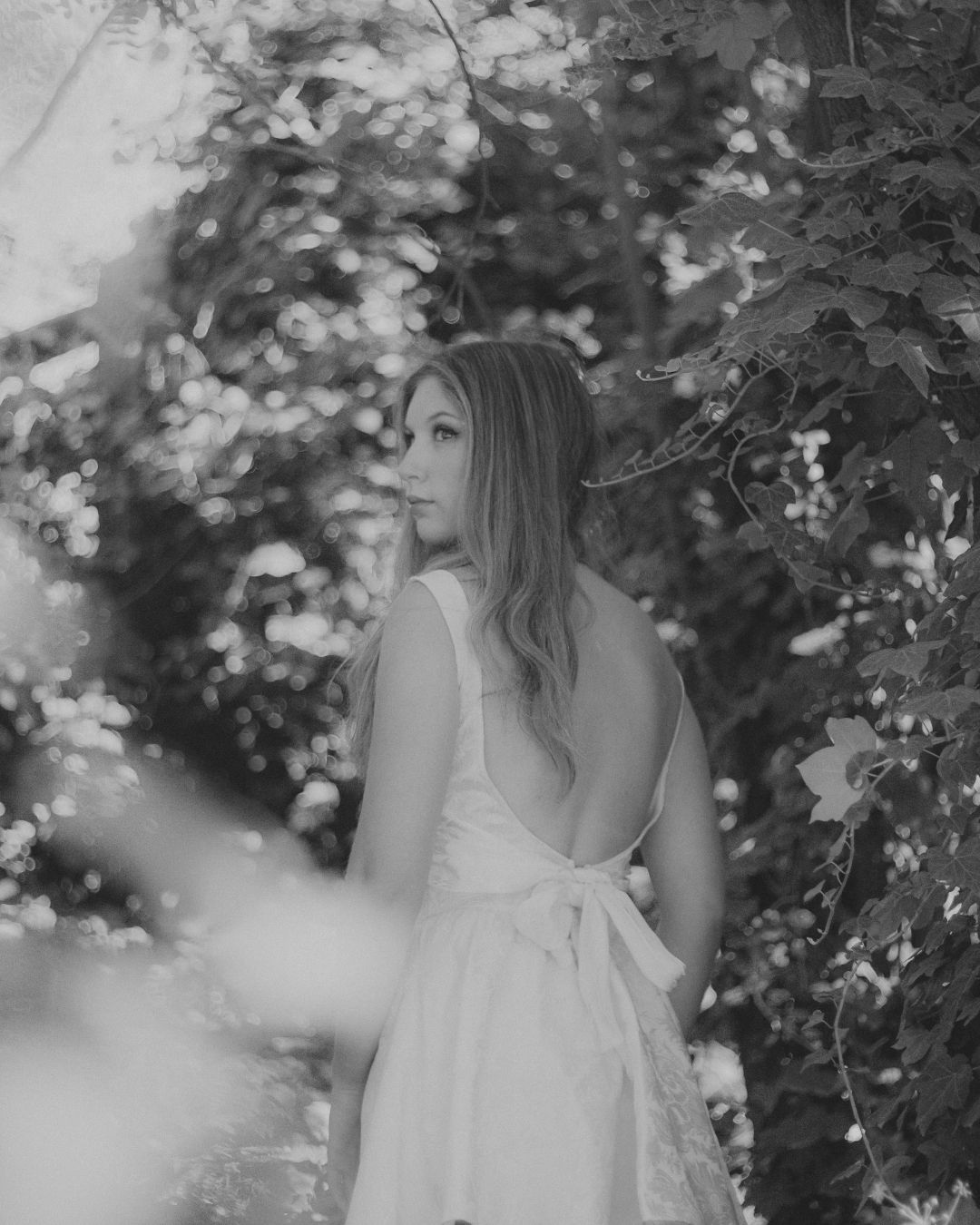 Black and white photo of woman in backless white dress standing among lush garden foliage.