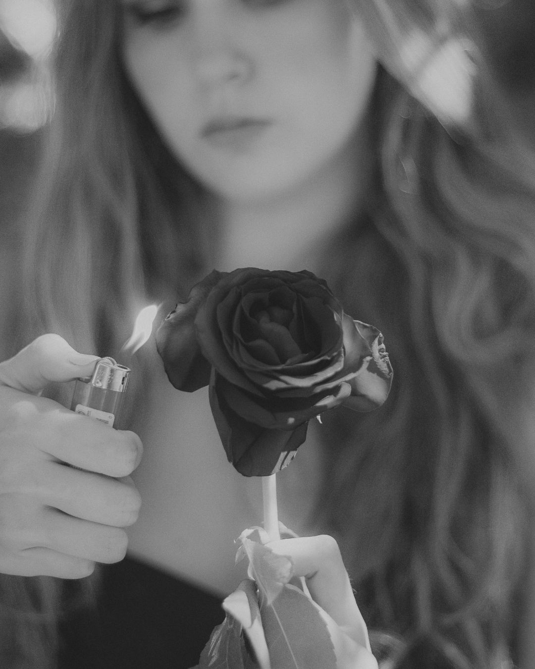 Black and white photo of a woman holding a dark rose near a lighter in a moody setting.