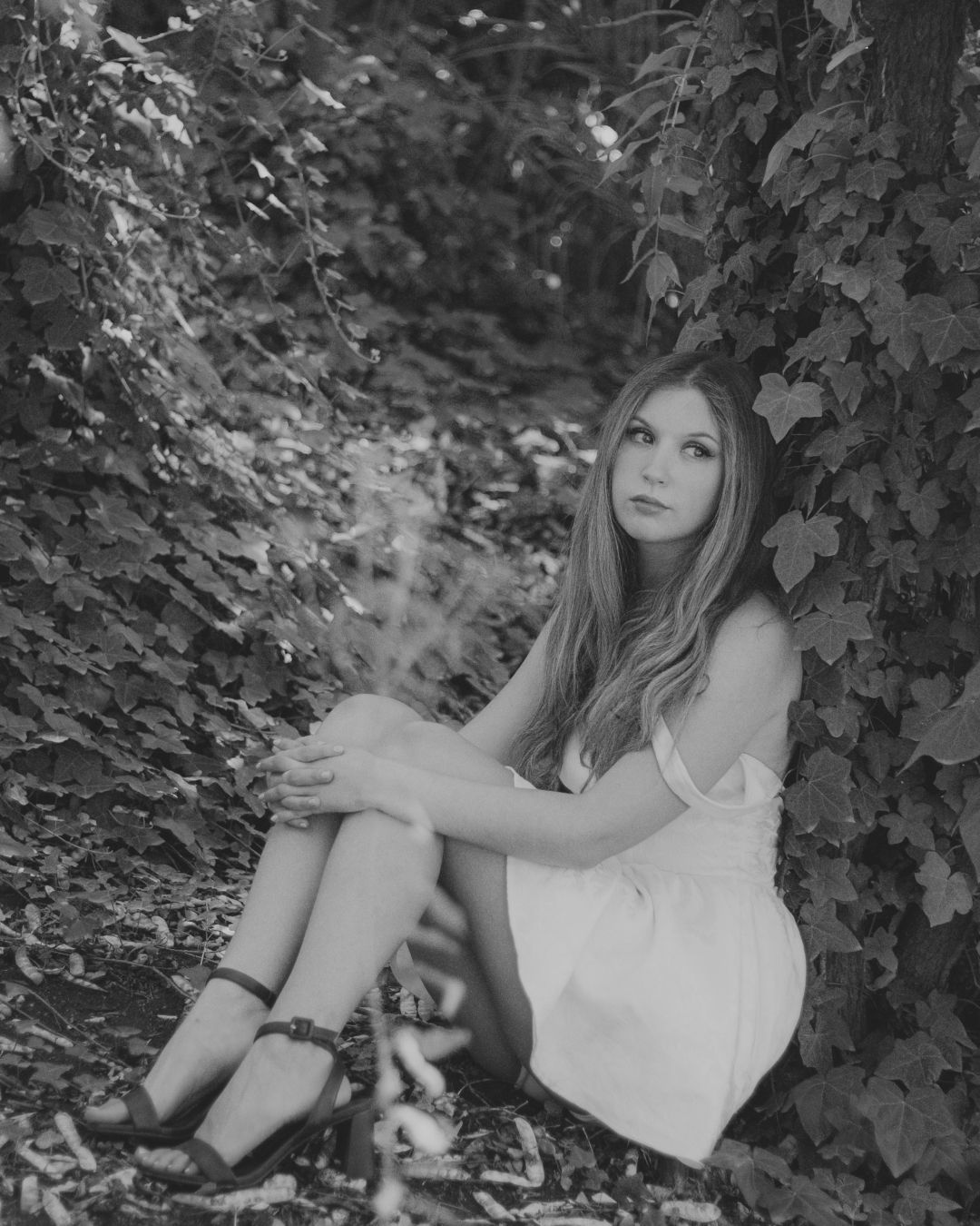 Black and white portrait of blonde woman in white dress sitting against ivy-covered wall.