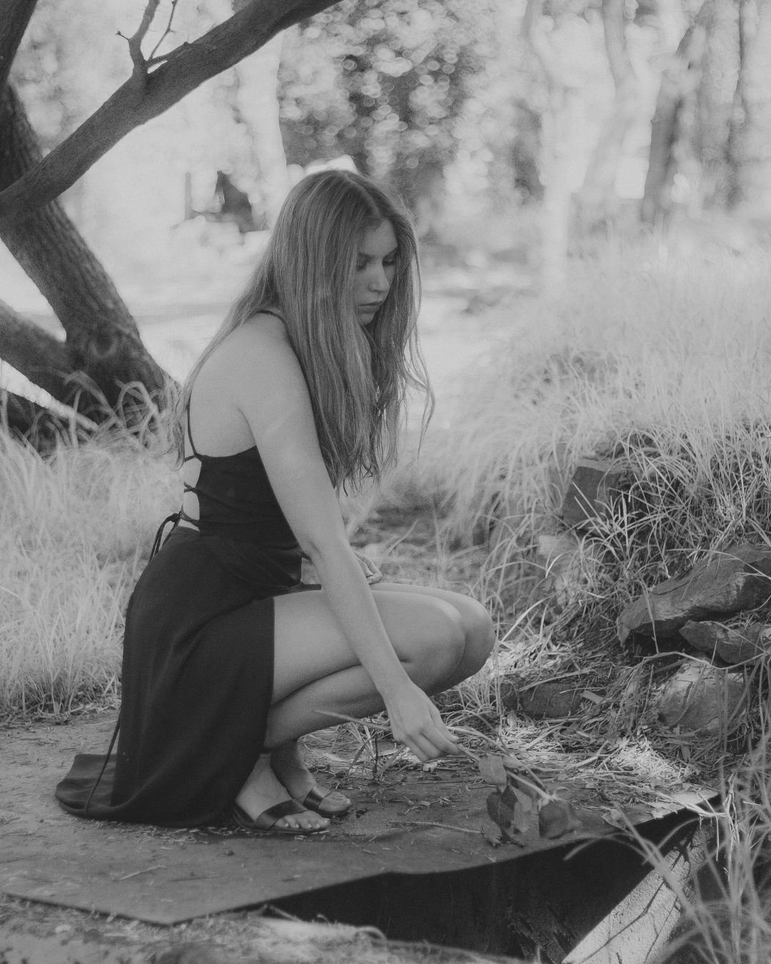 Black and white photo of blonde woman in black dress kneeling on dry grass outdoors.