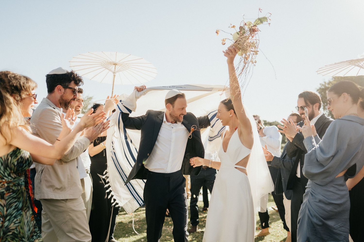 Joyful celebration outdoors with people throwing confetti and cheering under bright sunlight on a wedding day.
