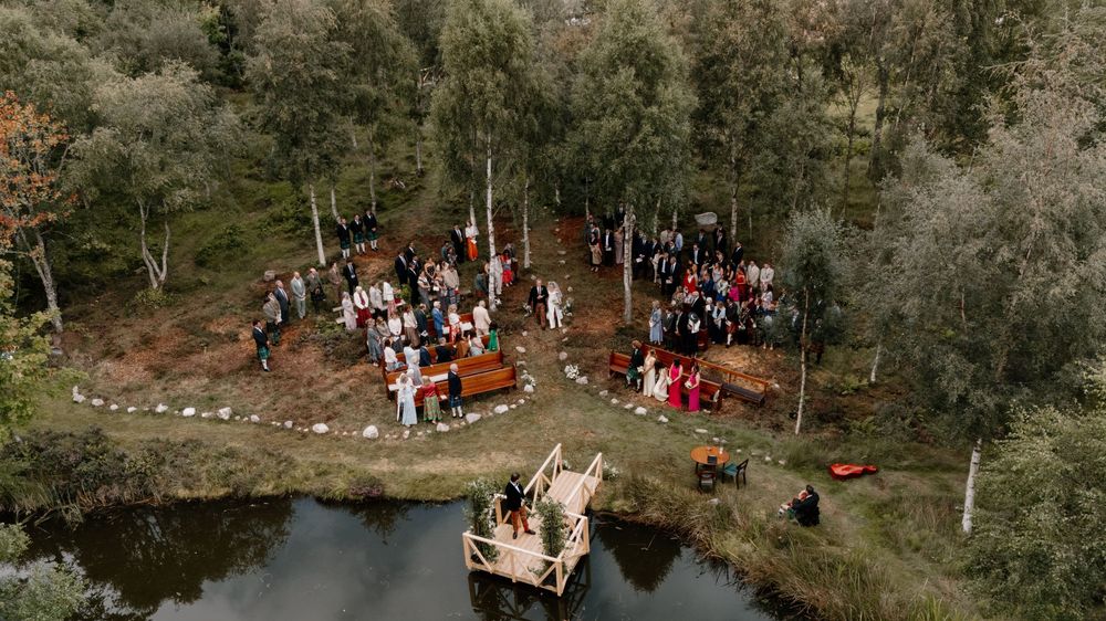 Aerial view of an outdoor woodland wedding ceremony with guests gathered among trees beside a small pond and wooden dock.
