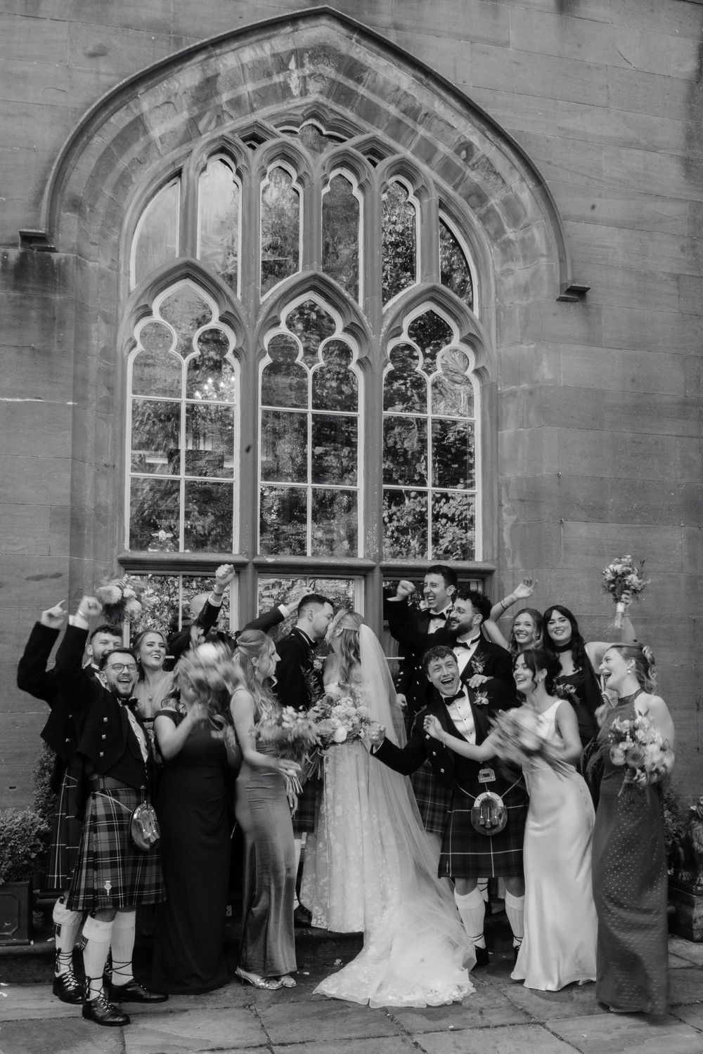 Black and white photo of a bride and groom celebrating with cheering wedding guests in kilts and gowns outside a Gothic arched window.