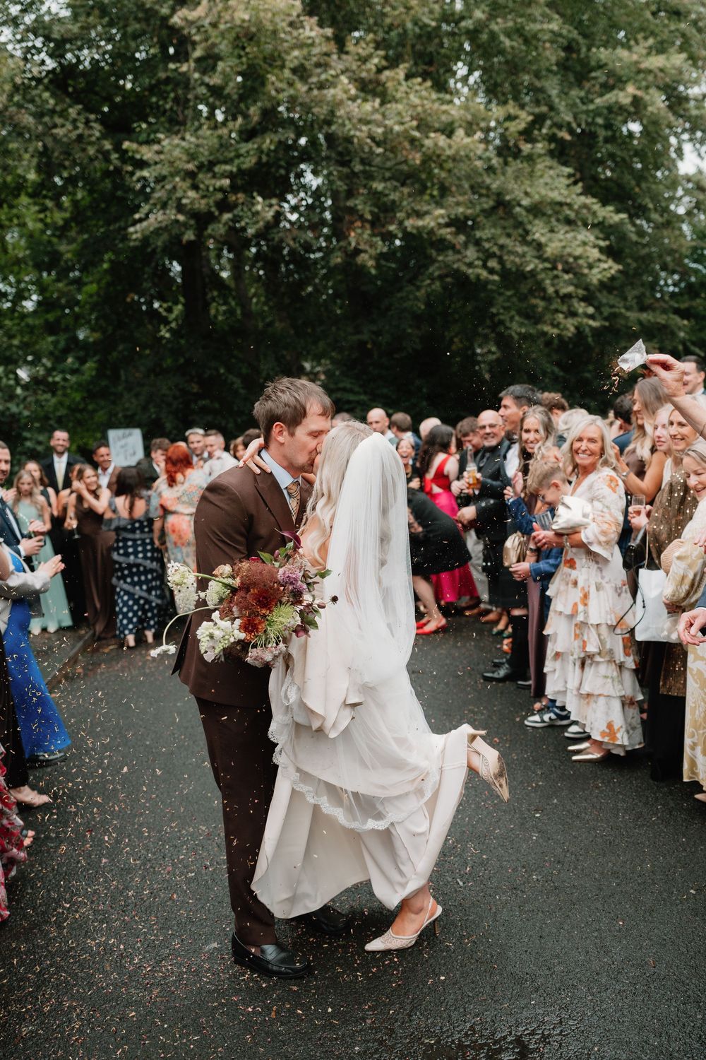Bride and groom kiss on a wet path as wedding guests cheer and throw confetti, with the bride holding a floral bouquet.