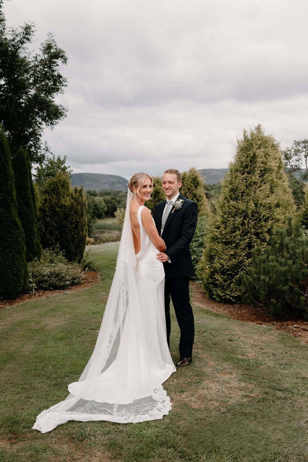 Bride in a elegant white gown with a long veil embraces her groom in a dark suit on a lush green lawn surrounded by evergreen trees.