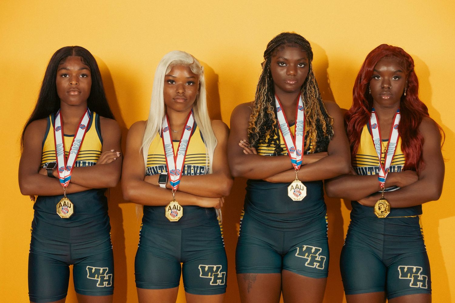 Four female track athletes with gold medals pose confidently against a yellow background.