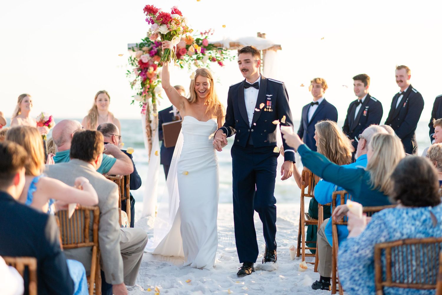 A person in a white wedding gown smiles while holding a vibrant bouquet of pink and red flowers outdoors.