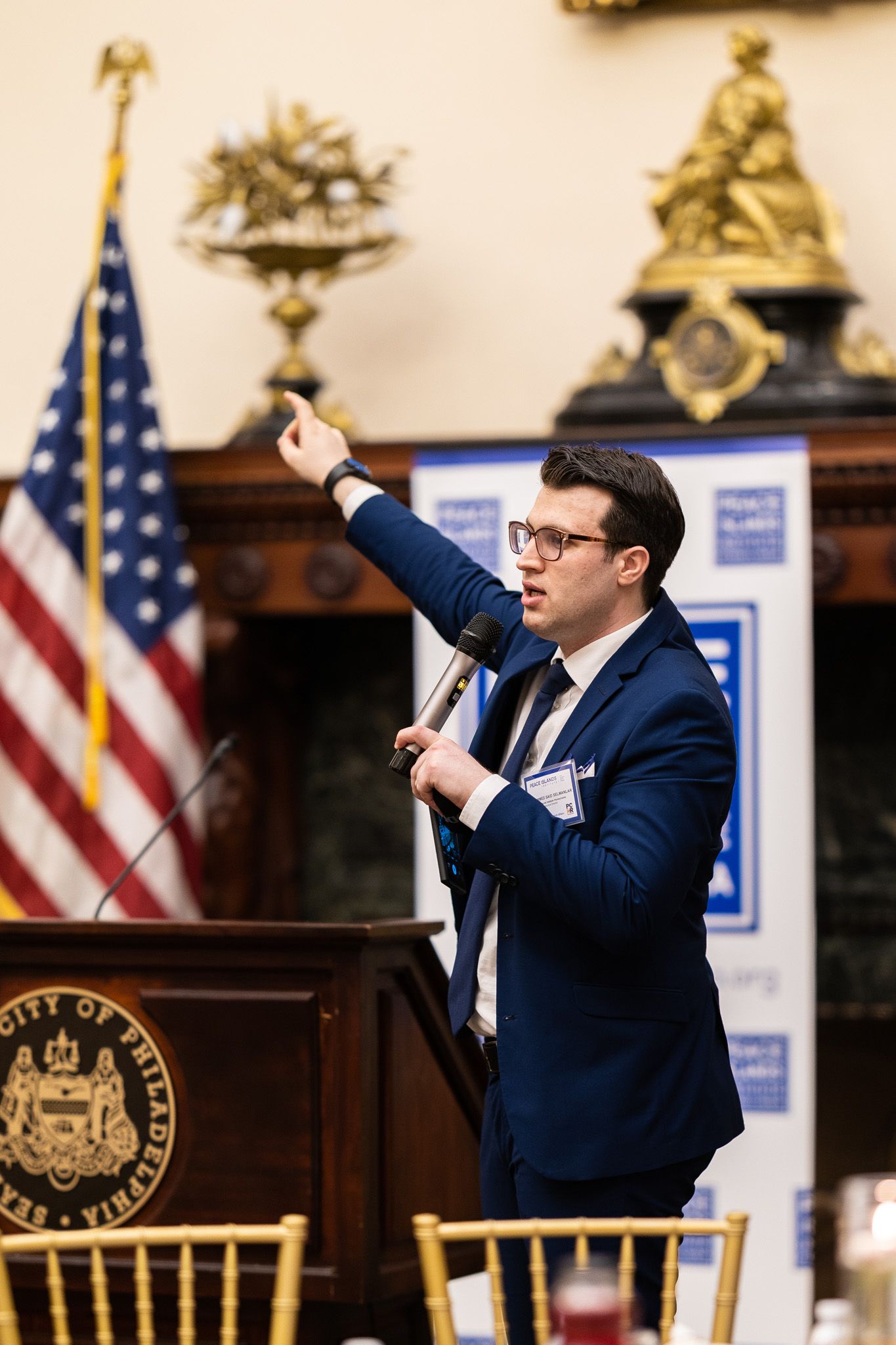 Speaker in navy suit gestures enthusiastically at podium with American flag in background.