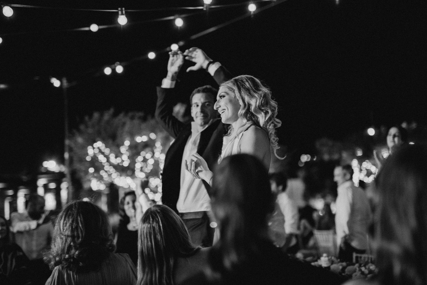 Black and white photo of festive crowd enjoying nighttime outdoor event with string lights overhead and raised arms in celebration.