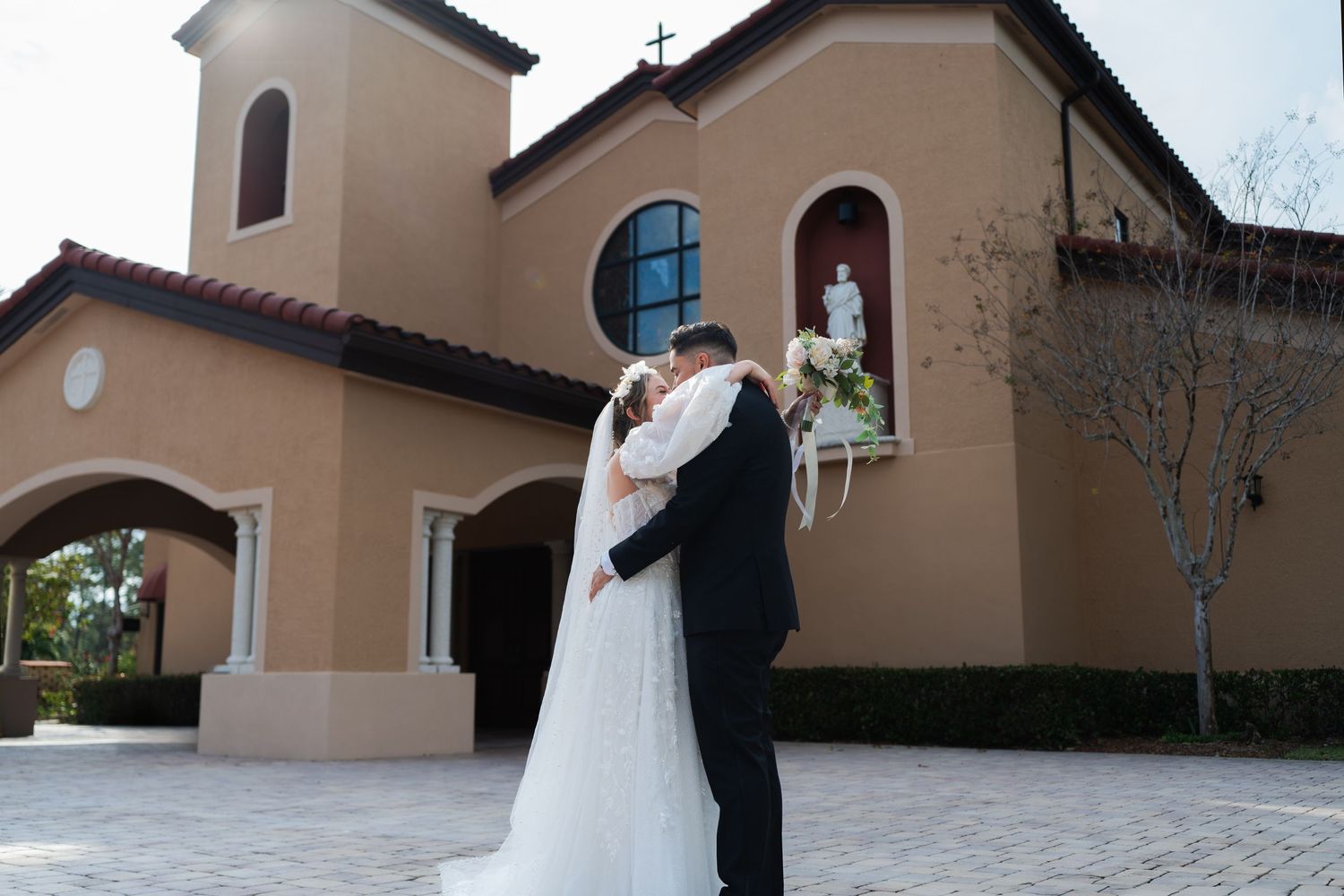 A couple in wedding attire embraces outside a Spanish-style church with stucco walls and arched architecture. Our Lady Queen of Apostles