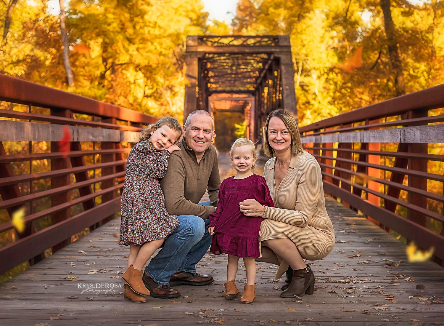 Family in fall attire poses on rustic wooden bridge surrounded by golden autumn foliage at sunset.
