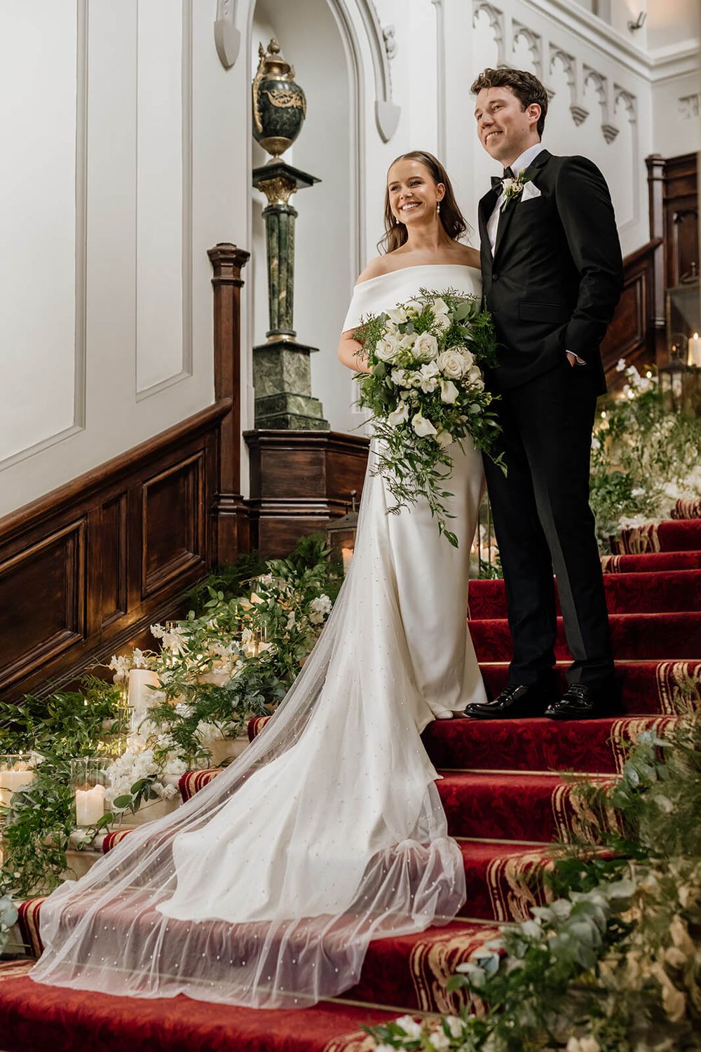 Wedding couple stands on red-carpeted staircase decorated with greenery and white flowers photographed by Dina Ozola Photography.