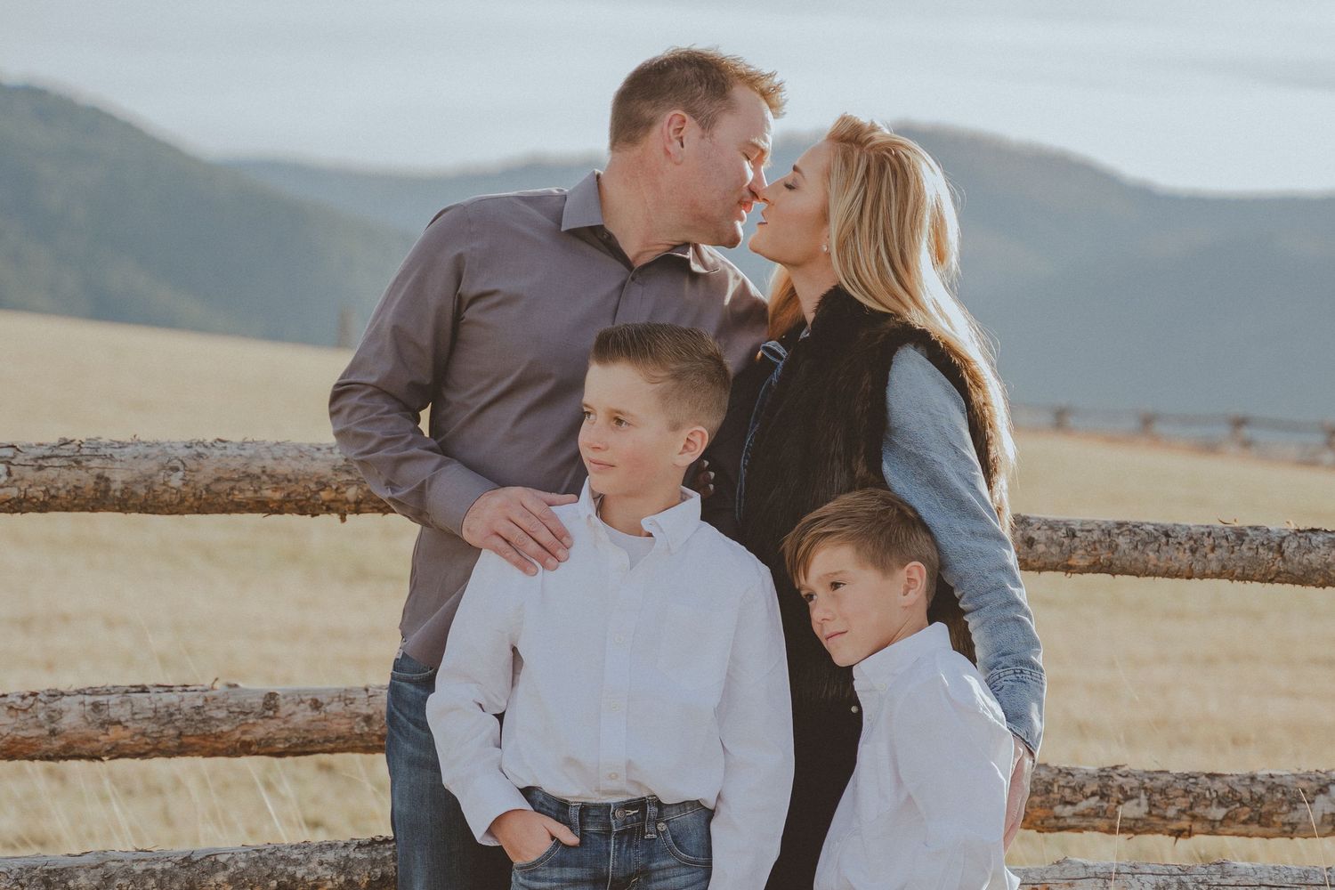 Family portrait against rustic wooden fence with mountains and fields in background.