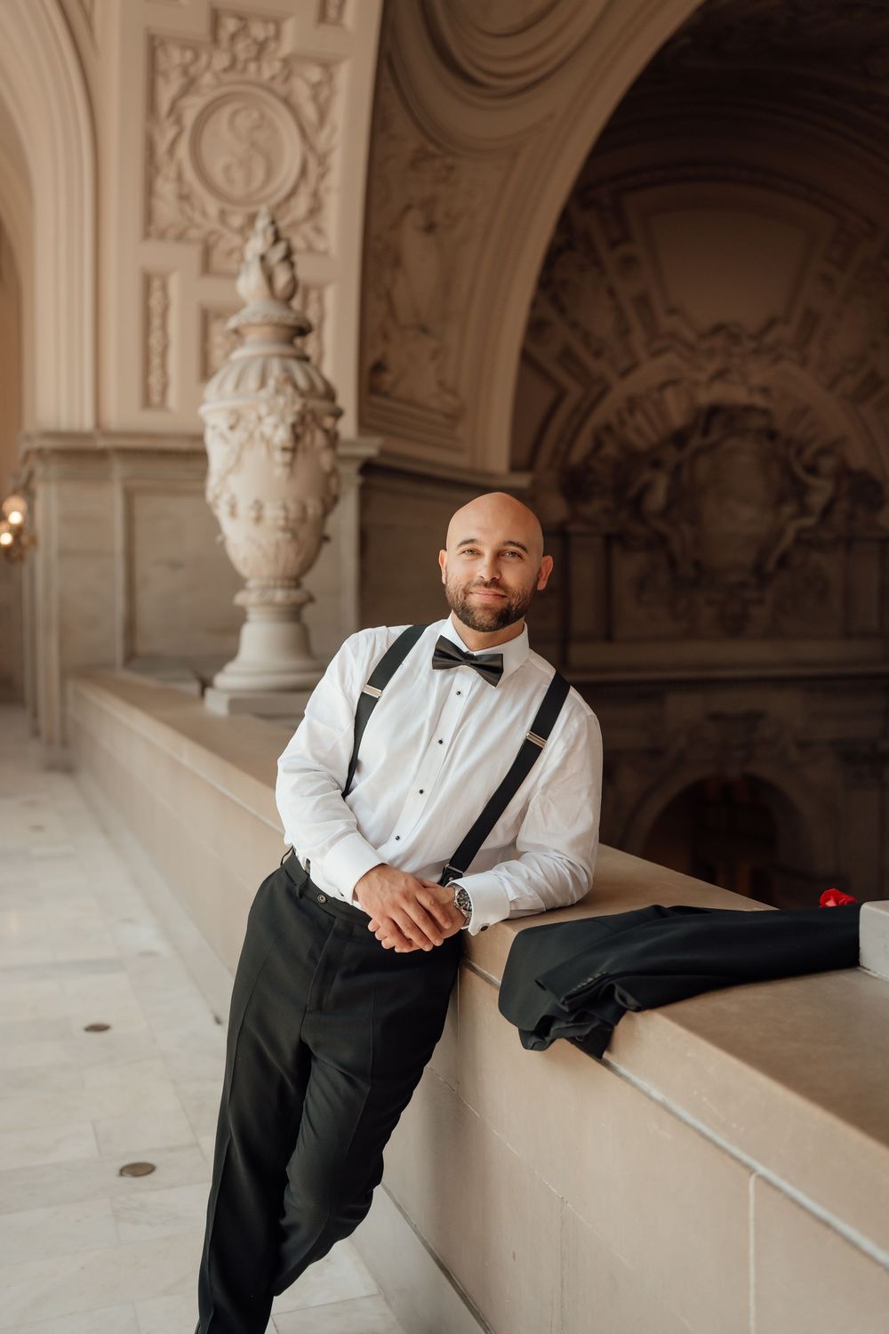 A groom in formal attire sits thoughtfully on a ledge in an ornate classical building with arched architecture.