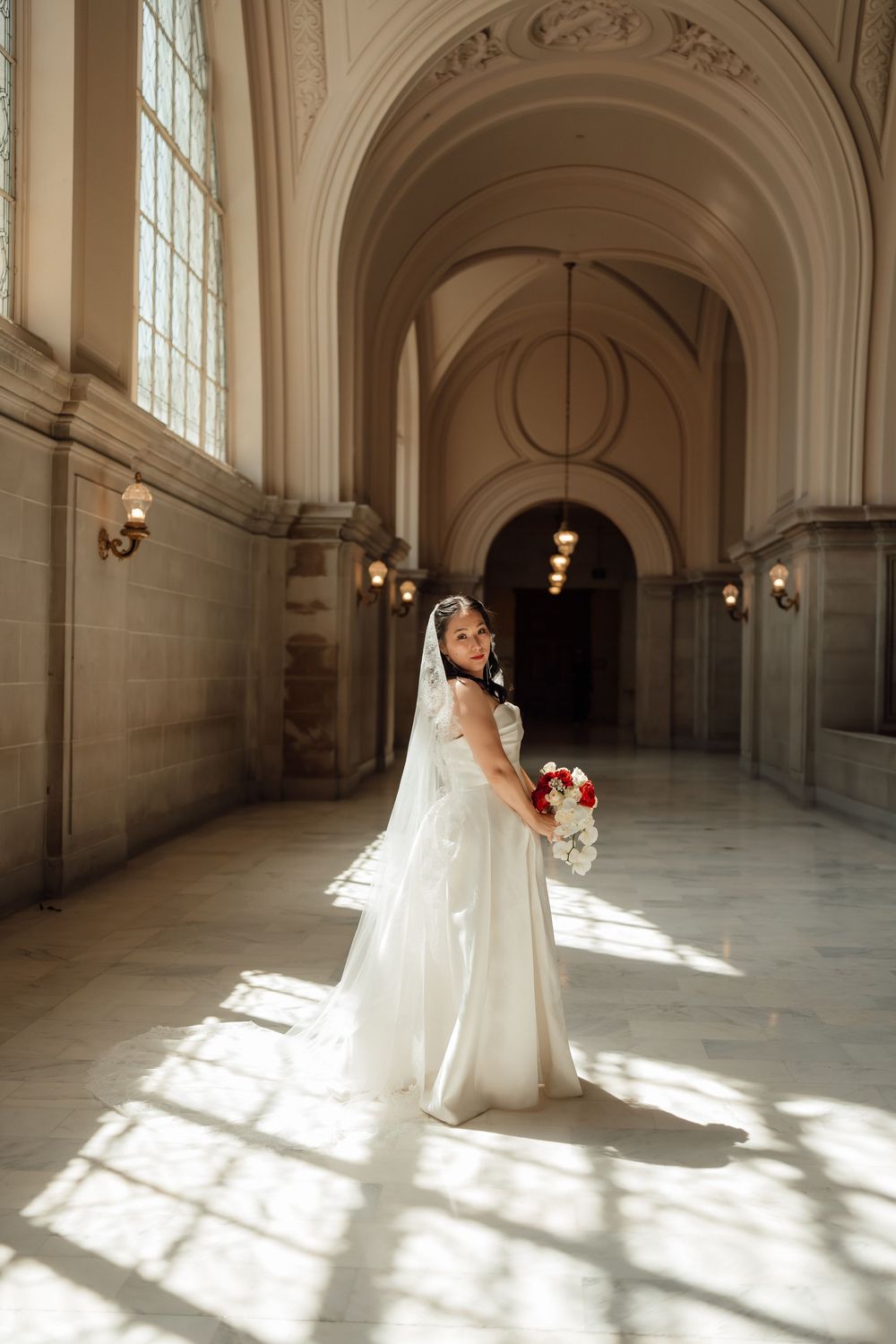 A bride in a flowing white gown and veil poses gracefully in a grand hallway with arched windows casting dramatic light.