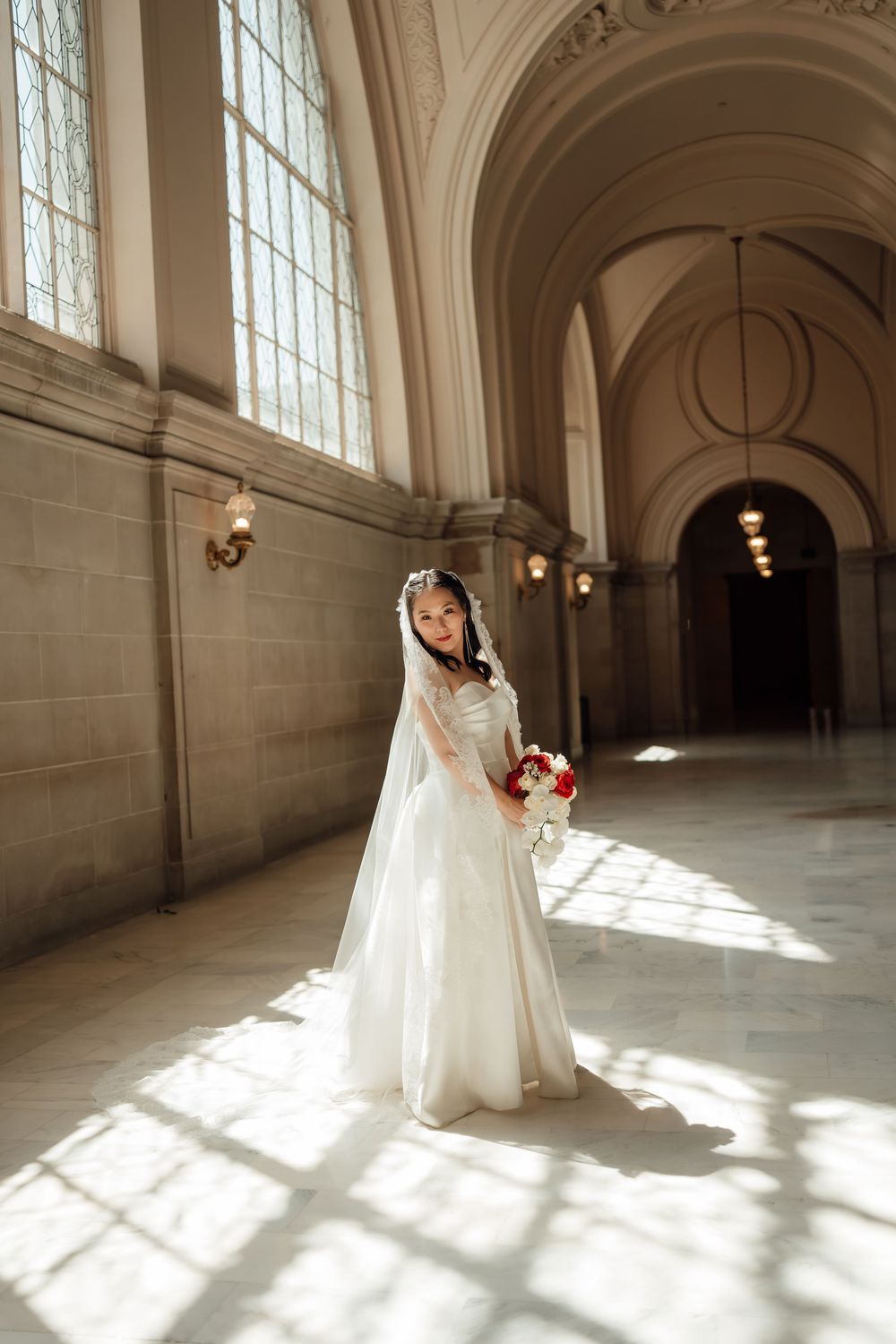 A bride in a flowing white gown and veil poses in an elegant hallway with arched ceilings and tall windows.