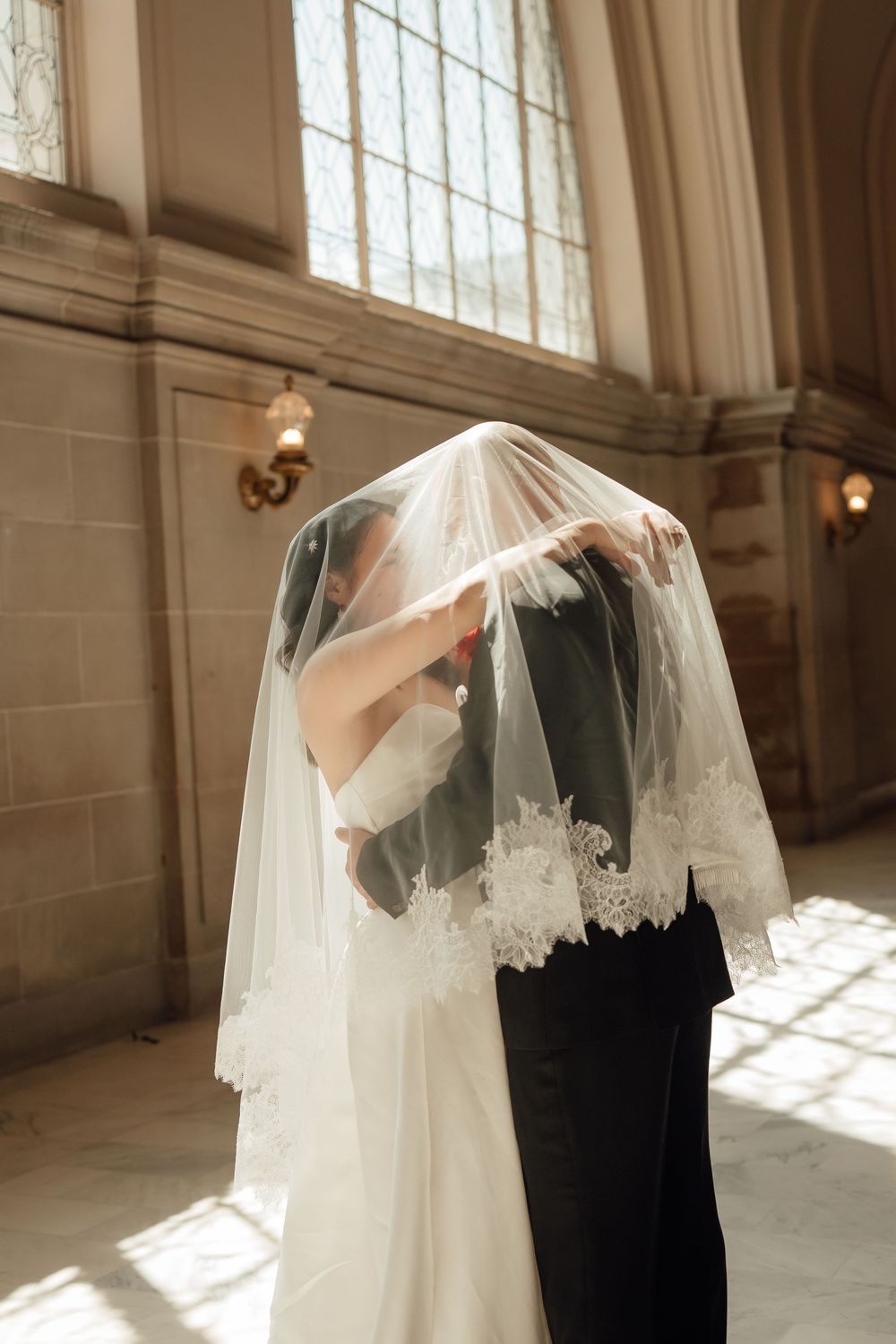 A romantic wedding couple shares a tender moment under a flowing white veil in a sunlit church interior.