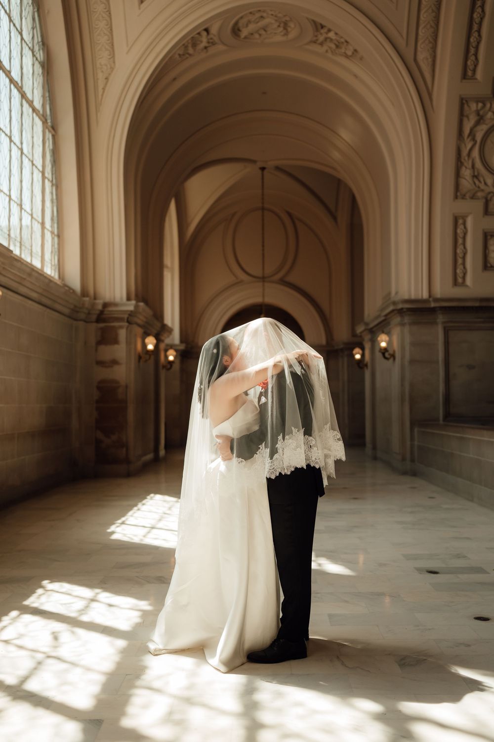 A couple shares an intimate moment under a flowing veil in a grand marble hall with arched windows.