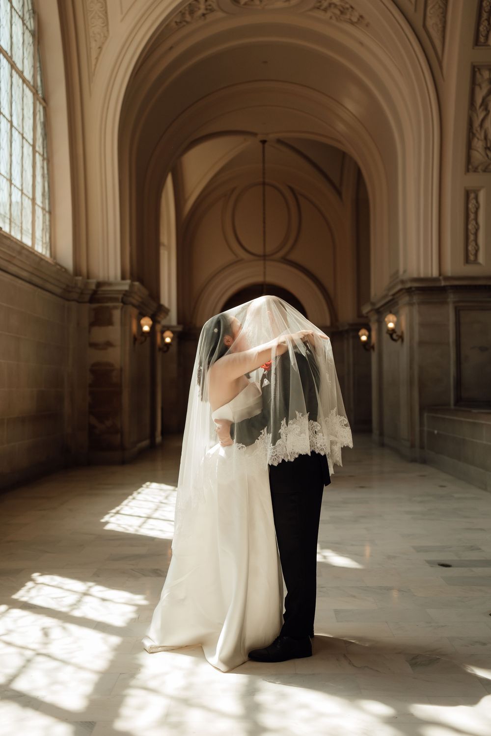 A romantic wedding moment captured in a sunlit hallway as the couple embraces under a delicate lace veil.