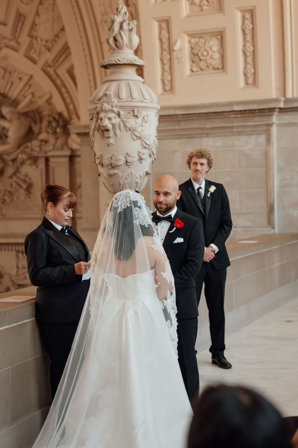A wedding ceremony takes place in an ornate hall with decorative architectural details and sculptures.